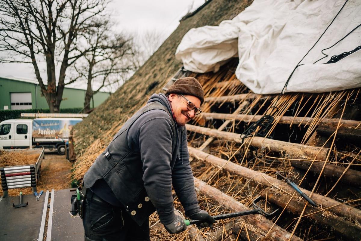 Ein Handwerker zwischen alten Rundholzlatten, darüber Planen, im Hintergrund Bäume.
