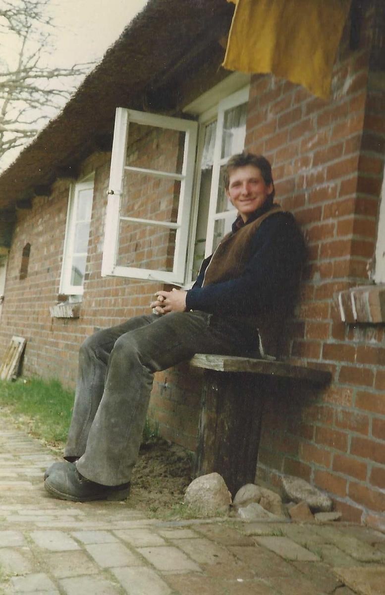 Ein Mann in Arbeitskleidung sitzt vor einem Backsteinhaus mit offenem Fenster.