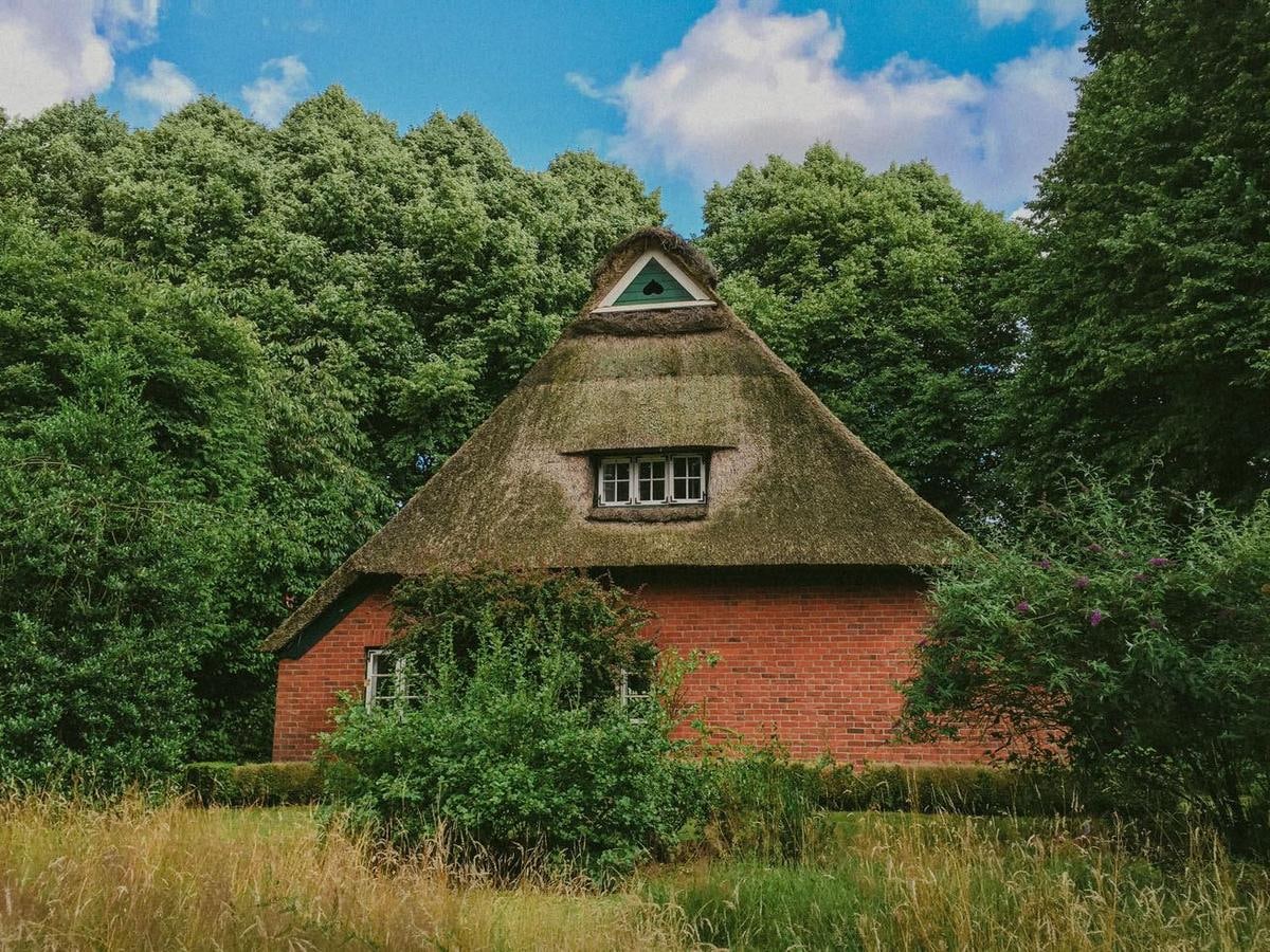 Kleines Backsteinhaus mit Reetdach und Gaube zwischen Bäumen.