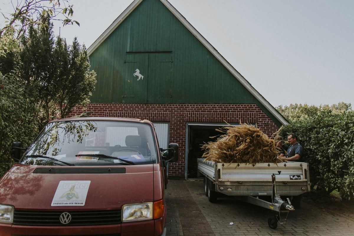 Roter Kleintransporter mit Anhänger voller Reet vor einer Scheune.