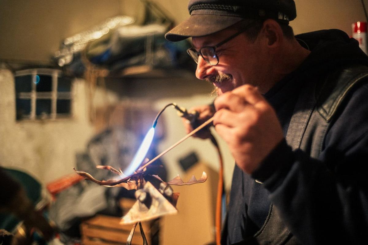 Ein Mann mit Mütze arbeitet in einer Werkstatt mit einem Lötbrenner an einem Metallteil.
