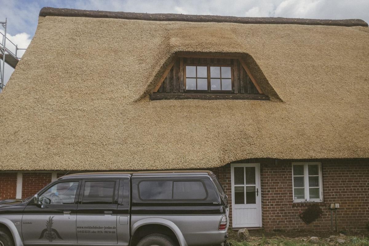 Hell leuchtendes, neues Reetdach mit Gaube über einem Backsteinhaus, davor ein Pick-up.
