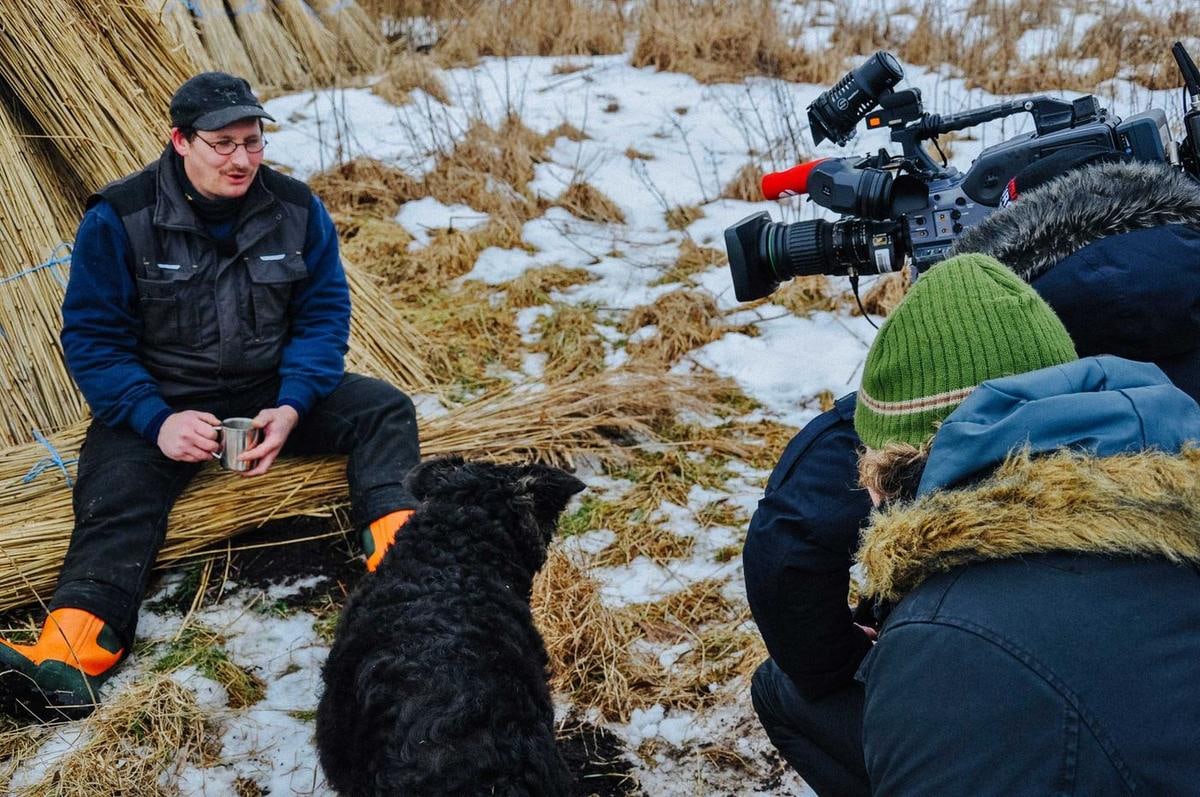 Ein Mann sitzt auf Reetbunden, daneben ein Hund und ein Kamerateam.