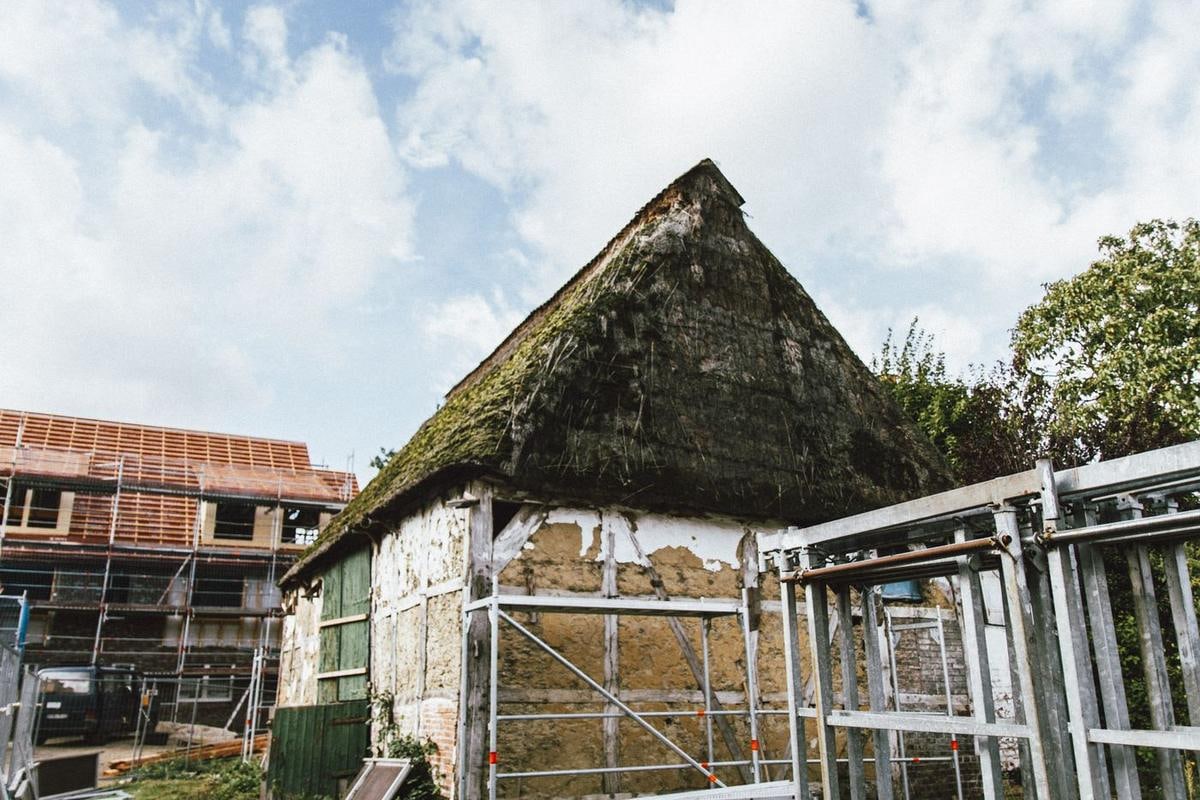 Alte Fachwerkscheune mit grünem Dach aus Moos und Reet, daneben Neubauten und ein Bauzaun.