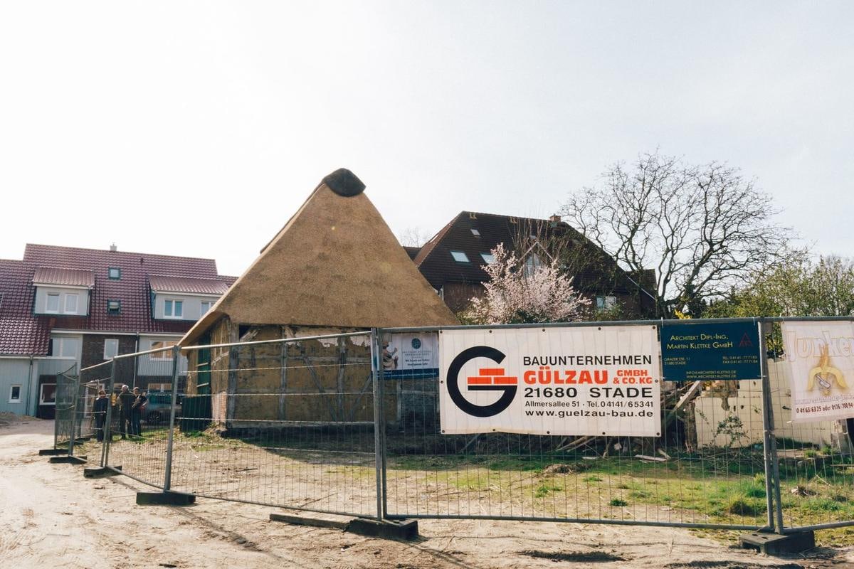 Scheune mit neuem Reetdach, davor Bauzaun mit Firmenschild.