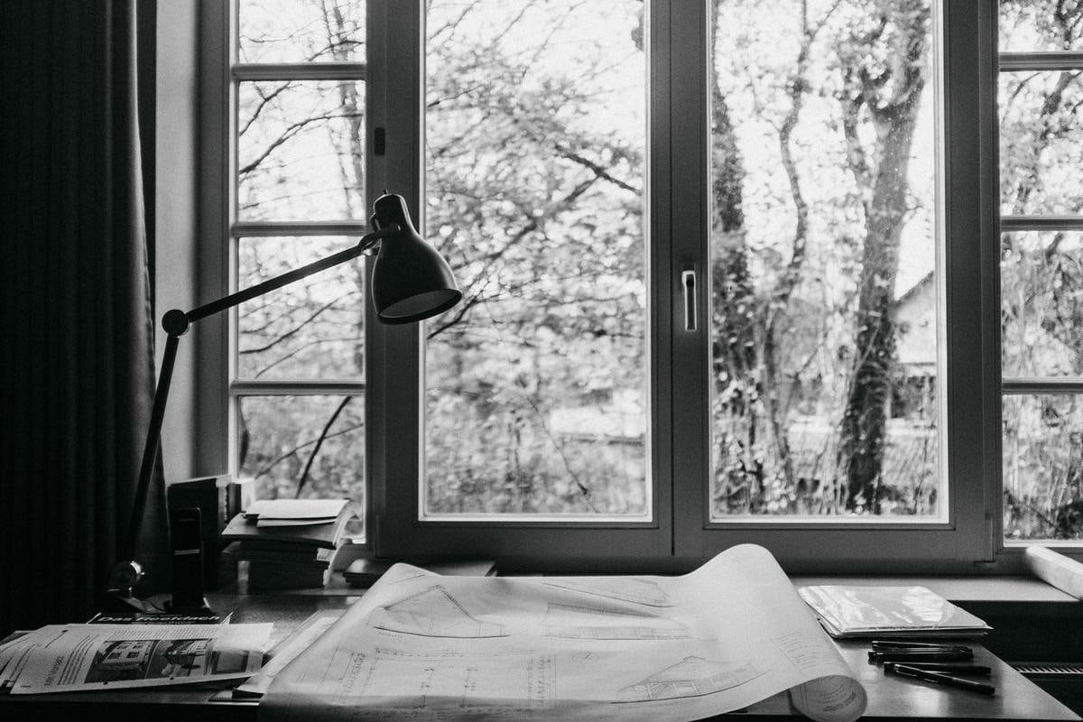 Schwarzweiß: Schreibtisch am Fenster, darauf ausgerollte Pläne.
