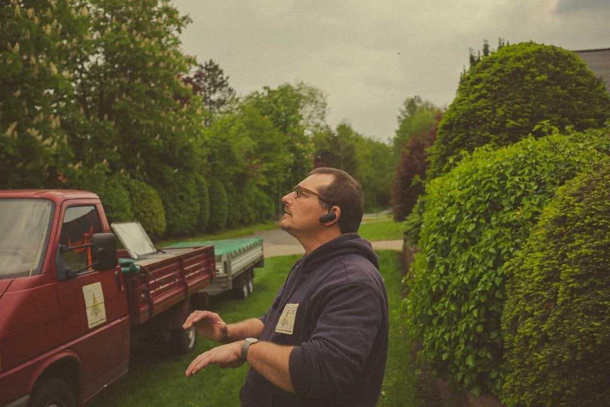 Ein Mann mit Mütze spricht neben einem roten Pick-up vor einer Hecke.