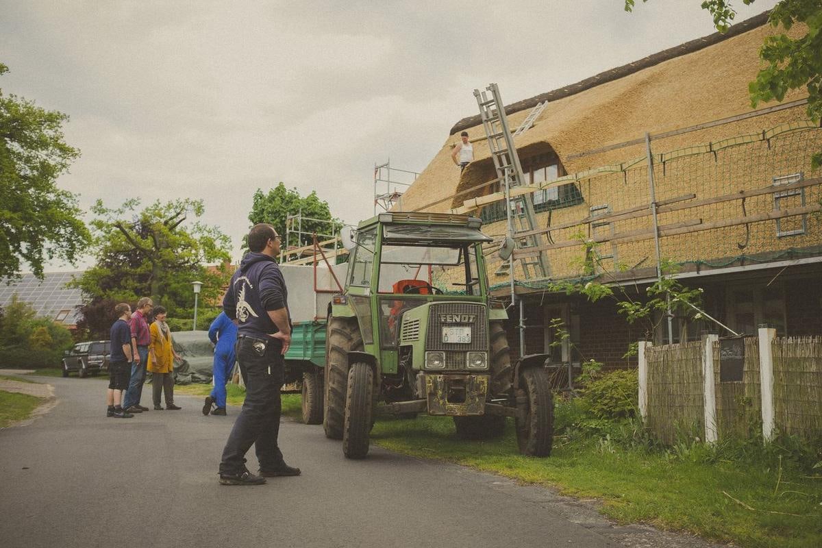 Grüner Trecker mit Anhänger vor einem eingerüsteten Reetdachhaus, Menschen auf der Straße.