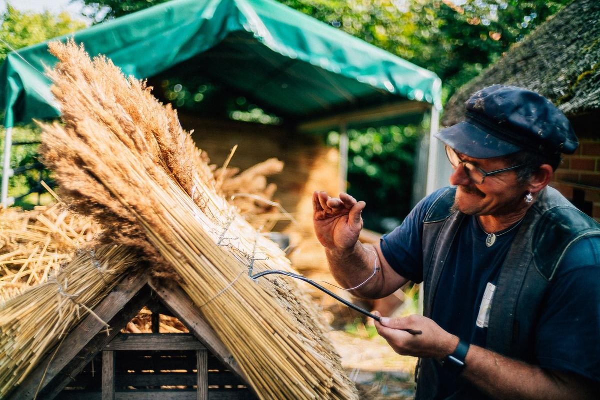 Ein Bund Reet liegt auf einem Vogelhaus, daneben arbeitet ein Mann.